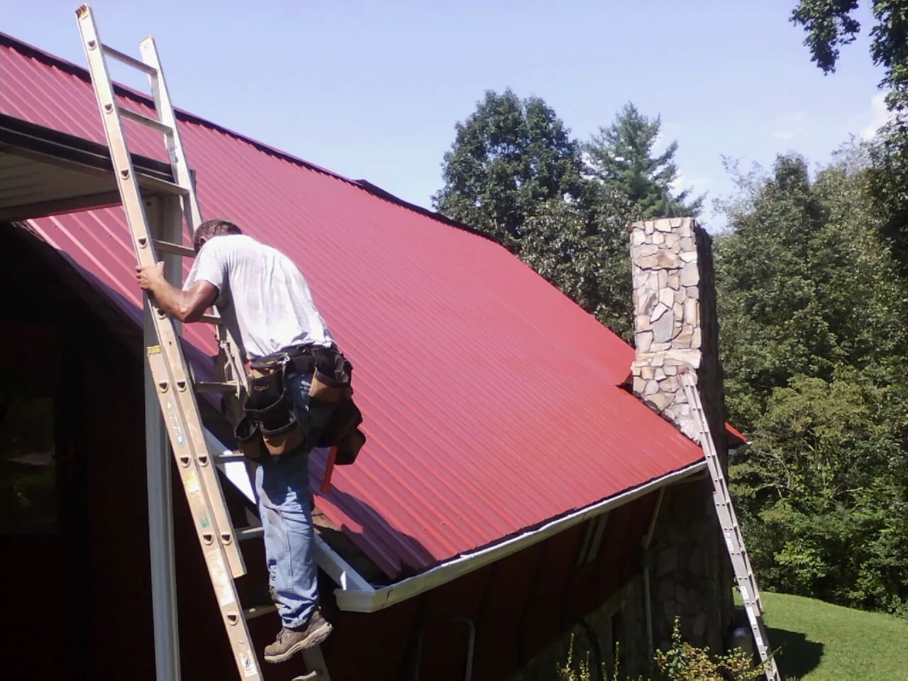 Roofer on a ladder beside a red metal roof and stone chimney — Soffit & Fascia Repair by Roofing Brothers in Eaton Rapids